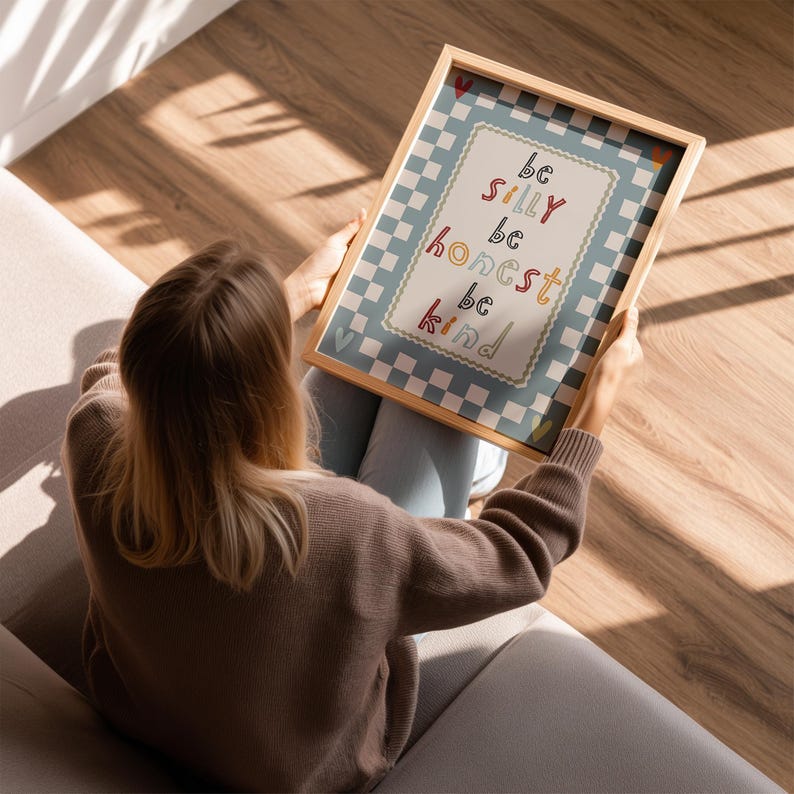 Pode incluir: Uma obra de arte emoldurada com as palavras "be silly, be honest, be kind" em letras coloridas. A obra de arte tem uma borda quadriculada azul clara e branca e uma moldura de madeira. A imagem &eacute; segurada por uma pessoa.