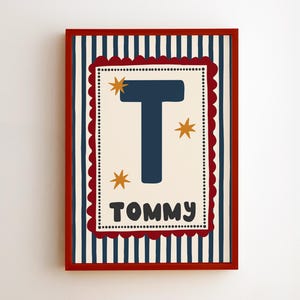Könnte beinhalten: Ein rechteckiger gerahmter Druck mit einem dunkelblauen Buchstaben T und dem Namen TOMMY in Dunkelgrau darunter. Das cremefarbene Rechteck in der Mitte hat einen gewellten roten Rand, goldene Sternakzente und einen marineblau-weiß gestreiften Hintergrund.
