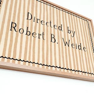May include: A framed sign with a light brown and white striped background. The text "Directed by Robert B. Weide" is printed in black. The frame is made of light-coloured wood, and the border has a black scalloped edge.