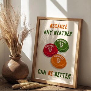 May include: A framed print with the text "BECAUSE ANY WEATHER CAN BE BETTER." The print features three colorful bottle caps with the Guinness logo. The frame is made of light brown wood, and the background is a neutral color.