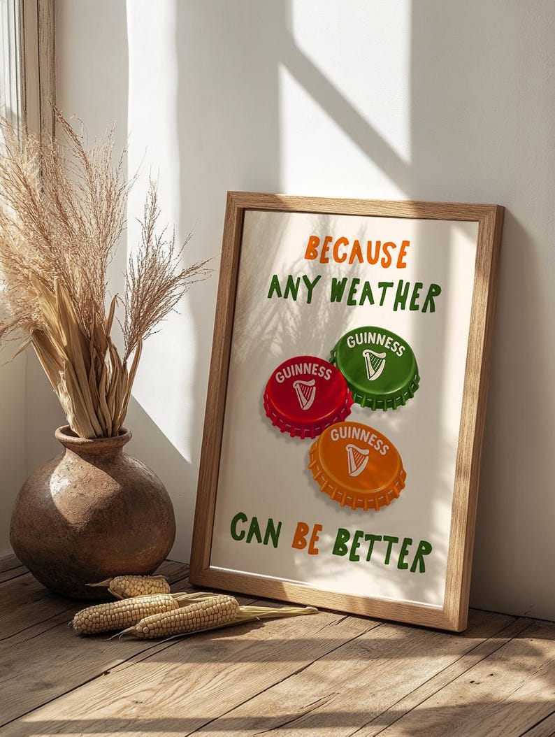 May include: A framed print with the text "BECAUSE ANY WEATHER CAN BE BETTER." The print features three colorful bottle caps with the Guinness logo. The frame is made of light brown wood, and the background is a neutral color.
