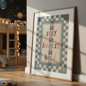 Pode incluir: Uma impress&atilde;o art&iacute;stica emoldurada com as palavras "be silly, be honest, be kind" em letras coloridas. A impress&atilde;o tem um fundo bege claro com uma borda quadriculada em azul claro e branco. A moldura &eacute; de cor de madeira clara.