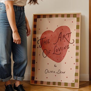 May include: A framed art print titled "The Art of Loving" by Olivia Dean. The artwork features a large red heart with the title in elegant script. The print has a pink background with red stars and a checkered border.