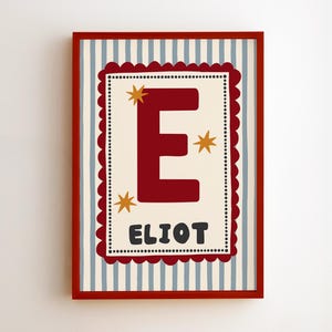 May include: A framed wall art print featuring a large dark red letter E and the name ELIOT in dark grey. The design includes three gold stars on a cream background, enclosed by a dark red scalloped border and a light blue and white striped frame.