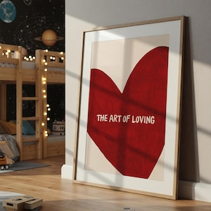 May include: Framed art print featuring a large red heart shape against a cream background. The text "THE ART OF LOVING" is centered within the heart. The artwork is displayed in a light wood frame.