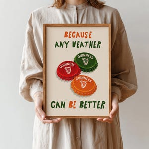 May include: A framed print featuring the text "BECAUSE ANY WEATHER CAN BE BETTER". The print includes three Guinness bottle caps in red, green, and orange. The Guinness logo is visible on each cap. The background is a light beige color.