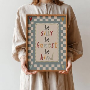 Pode incluir: Uma impress&atilde;o art&iacute;stica emoldurada com as palavras "be silly, be honest, be kind" em letras coloridas. A impress&atilde;o tem uma borda xadrez azul claro e branco. Uma pessoa segura a impress&atilde;o.