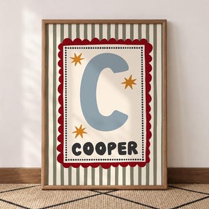 May include: A framed wall art print featuring a large light blue letter C and the name COOPER in black text. The design includes three yellow stars on a cream background, enclosed by a scalloped red border and a dotted line, set on striped green.