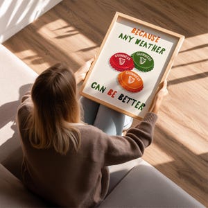 May include: A framed print featuring the text "BECAUSE ANY WEATHER CAN BE BETTER" with three Guinness bottle cap designs in red, green, and orange. The print is held by a person sitting on a couch.
