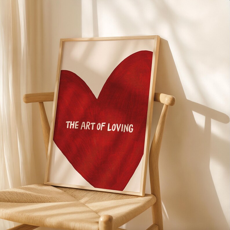 May include: A framed art print featuring a large red heart on a cream background. The words "THE ART OF LOVING" are printed in white across the bottom of the heart. The artwork is displayed on a wooden chair.