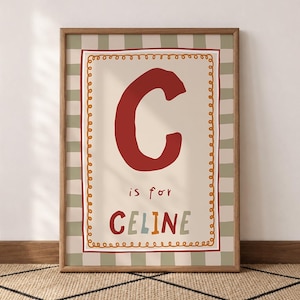 May include: A framed wall art print featuring a large dark red letter C, the text is for, and the name CELINE in multicoloured letters. The design includes a decorative orange scalloped border on a cream background with a sage green striped border.