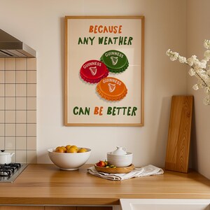 May include: A framed print with the text "BECAUSE ANY WEATHER CAN BE BETTER" and images of Guinness bottle caps. The caps are red, green, and orange. The print is displayed in a kitchen setting with a wooden countertop.