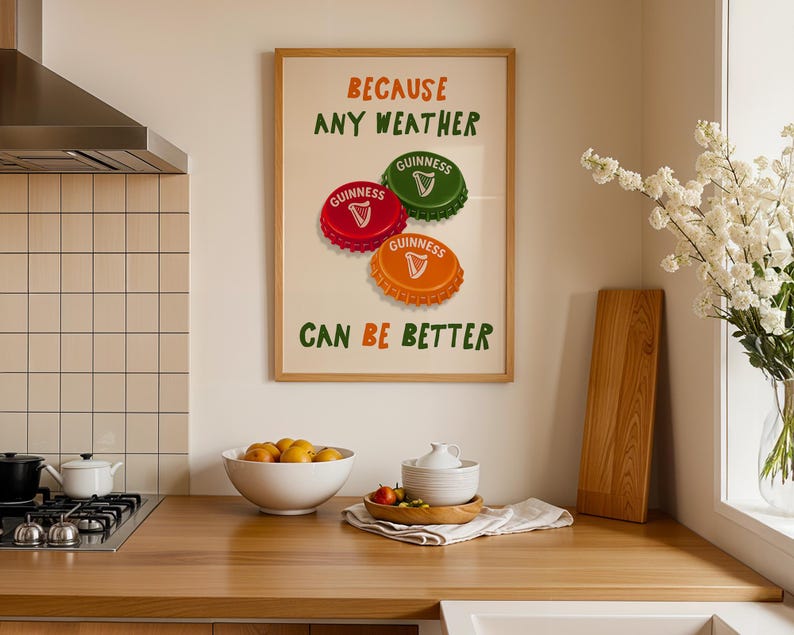 May include: A framed print with the text "BECAUSE ANY WEATHER CAN BE BETTER" and images of Guinness bottle caps. The caps are red, green, and orange. The print is displayed in a kitchen setting with a wooden countertop.
