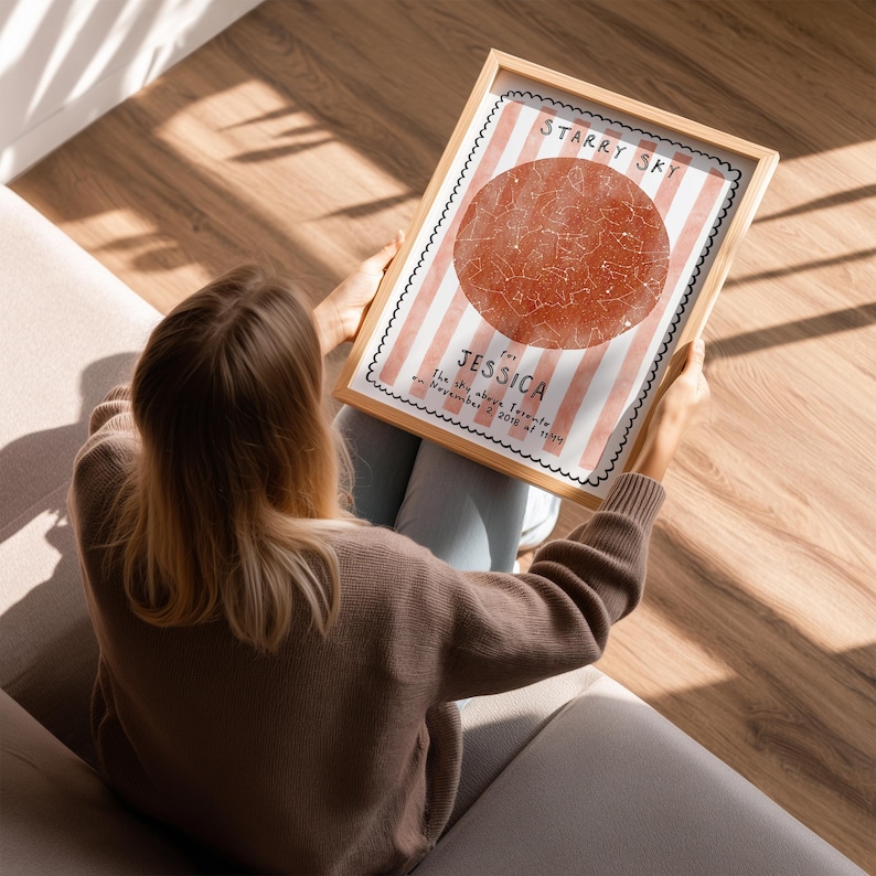 May include: A framed print with the text "STARRY SKY" and "JESSICA" over a red circle with a star map design. The background has pink and white stripes. The frame is light brown.