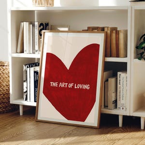 May include: A framed art print featuring a large red heart on a cream background. The text "THE ART OF LOVING" is centered within the heart. The print is displayed on a wooden floor, leaning against a white bookshelf filled with books.