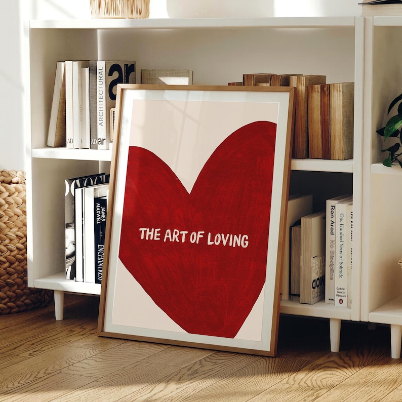 May include: A framed art print featuring a large red heart on a cream background. The text "THE ART OF LOVING" is centered within the heart. The print is displayed on a wooden floor, leaning against a white bookshelf filled with books.