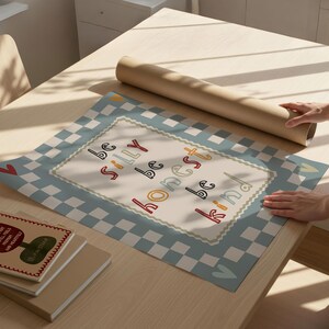 Pode incluir: Uma mesa de madeira clara cont&eacute;m um rolo de papel castanho, uma pilha de livros e um tapete decorativo. O tapete apresenta uma borda xadrez azul e branca e as palavras "be silly be honest be kind".