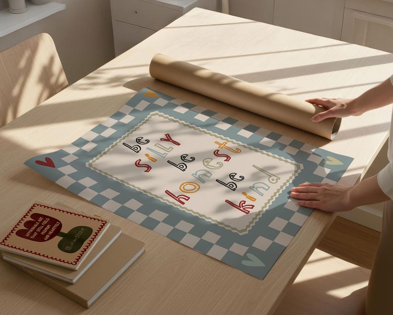 Pode incluir: Uma mesa de madeira clara cont&eacute;m um rolo de papel castanho, uma pilha de livros e um tapete decorativo. O tapete apresenta uma borda xadrez azul e branca e as palavras "be silly be honest be kind".