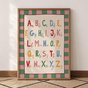 May include: A framed alphabet poster featuring uppercase and lowercase letters in various colors on a cream background. The border consists of alternating vertical green and pink stripes, enclosed by a thin red line, all within a wooden frame.