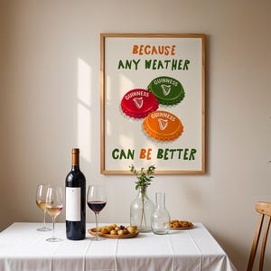 May include: A framed print with the text "BECAUSE ANY WEATHER CAN BE BETTER" and three Guinness bottle caps in red, green, and orange. A bottle of wine, wine glasses, and snacks are on a white tablecloth.