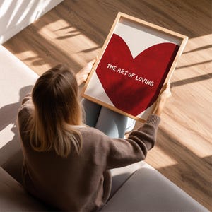 May include: A framed print featuring a large red heart on a cream background. The text "THE ART OF LOVING" is displayed in white. The frame is made of light-colored wood. The print is held by a person.