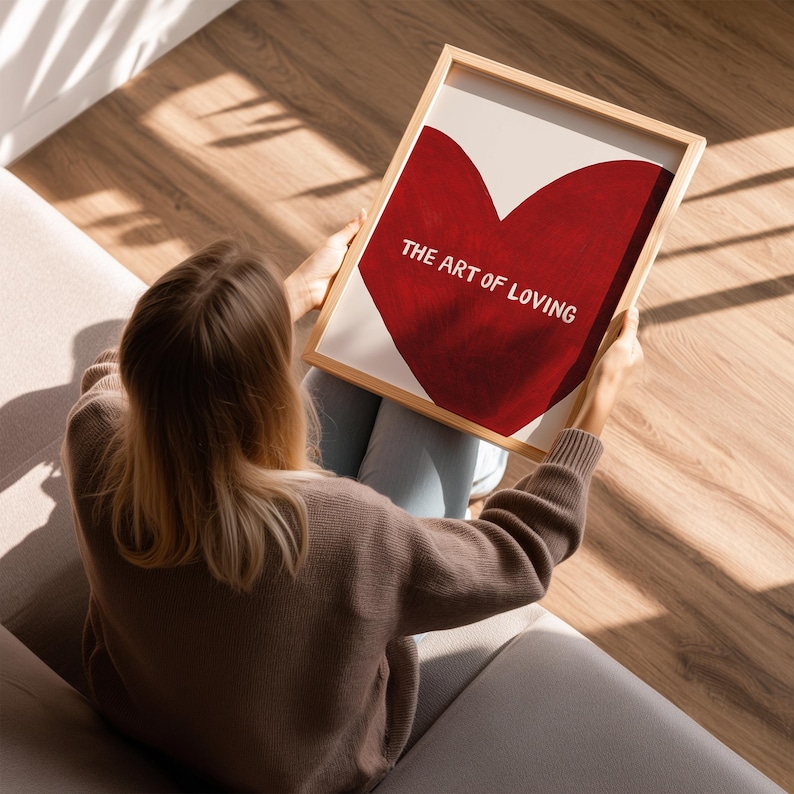 May include: A framed print featuring a large red heart on a cream background. The text "THE ART OF LOVING" is displayed in white. The frame is made of light-colored wood. The print is held by a person.