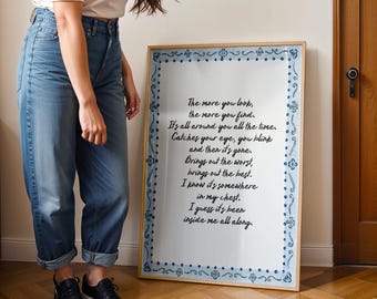 May include: A framed print with a light wood frame and a white background. The print has a blue decorative border and black handwritten text that reads, "The more you look, the more you find..." The print is displayed indoors.