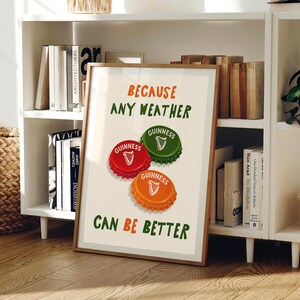 May include: A framed print featuring three Guinness bottle caps in red, green, and orange, with the Guinness logo. The text reads "BECAUSE ANY WEATHER CAN BE BETTER" in green and orange. The print is displayed on a white shelf.