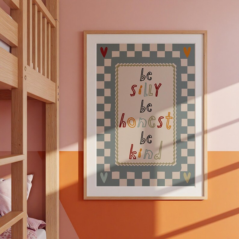 Pode incluir: Uma impress&atilde;o art&iacute;stica emoldurada com as palavras "be silly, be honest, be kind" em letras coloridas. A impress&atilde;o tem uma borda quadriculada e &eacute; exibida em uma parede rosa e laranja. Uma estrutura de beliche de madeira &eacute; vis&iacute;vel.