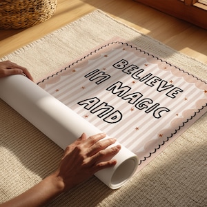 May include: A poster with the words "BELIEVE IN MAGIC AND" printed on it, being unrolled. The poster has a pink and white striped background with star accents and a scalloped border. The poster is on a beige rug.