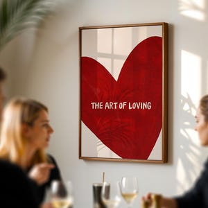 May include: A framed art print featuring a large red heart with the words "THE ART OF LOVING" in white. The artwork is displayed on a white background and framed in a wooden frame. The image is hanging on a wall.