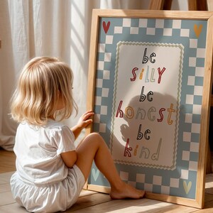 Pode incluir: Uma impress&atilde;o emoldurada com as palavras "be silly, be honest, be kind" em letras coloridas. A impress&atilde;o tem uma borda xadrez azul clara e est&aacute; apoiada em um cavalete de madeira. Uma crian&ccedil;a pequena est&aacute; sentada ao lado da impress&atilde;o.