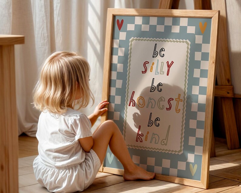 Pode incluir: Uma impress&atilde;o emoldurada com as palavras "be silly, be honest, be kind" em letras coloridas. A impress&atilde;o tem uma borda xadrez azul clara e est&aacute; apoiada em um cavalete de madeira. Uma crian&ccedil;a pequena est&aacute; sentada ao lado da impress&atilde;o.