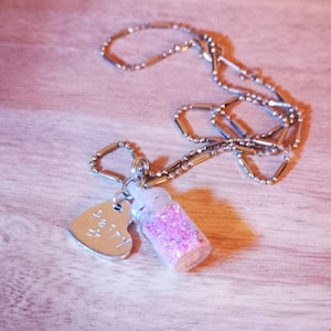 May include: A silver necklace with a small glass bottle charm filled with pink glitter and a silver heart charm inscribed with the word "fairy".