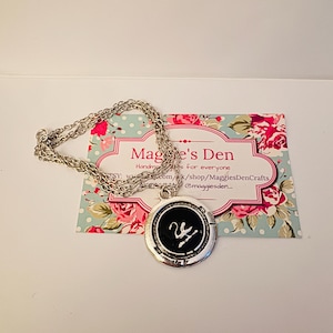 May include: A silver-toned necklace with a circular pendant featuring a black center and a white swan design. The pendant is on a silver chain, resting on a floral-patterned card with the text "Maggie's Den."