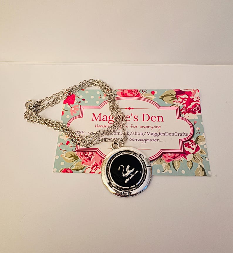 Puede incluir: Un collar plateado con un colgante circular con un centro negro y un dise&ntilde;o de cisne blanco. El colgante est&aacute; sobre una tarjeta con estampado floral con el texto "Maggie's Den".