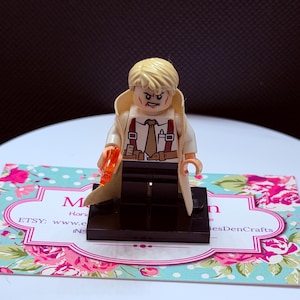 May include: A blonde LEGO minifigure wearing a tan trench coat over a white shirt with a brown tie and red suspenders. The figure has a determined facial expression with red markings, black trousers, and holds a small translucent orange accessory.