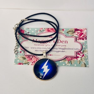 May include: A round pendant necklace with a white lightning bolt design on a dark blue background. The pendant is set in a silver-colored metal frame and hangs from a black cord necklace. The necklace rests on a floral patterned card with the text "Maggie's Den".