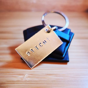 Op de afbeelding: Zilveren sleutelhanger met een rechthoekig label waarop in hoofdletters "BITCH!" staat. De sleutelhanger is bevestigd aan een zwarte leren sleutelhanger.