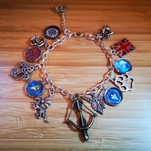 Puede incluir: Un brazalete de plata con una variedad de dijes, que incluyen un dragón, un arco y una flecha, una flor de lis, una huella de pata, un hada, una corona, una rosa, una brújula, una bandera y un símbolo.