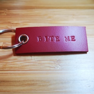 Könnte beinhalten: Ein roter Leder-Schlüsselanhänger mit dem geprägten Schriftzug "BITE ME". Er hat einen silbernen Metallring zur Befestigung von Schlüsseln. Der Hintergrund ist eine hellbraune Holzoberfläche.