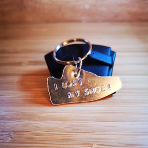 Könnte beinhalten: Ein goldfarbener schuhförmiger Schlüsselanhänger mit dem eingravierten Text "Ich habe meinen Schuh verloren!" Der Schlüsselanhänger ist an einem schwarzen Leder-Schlüsselanhänger befestigt.