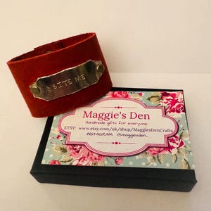 Könnte beinhalten: Ein rotes Lederarmband mit dem Schriftzug "BITE ME" auf einem Metallschild. Daneben eine schwarze Schachtel mit floralem Muster und dem Text "Maggie's Den".