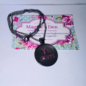 May include: A black pendant necklace with a round pendant that has the words "YA IDJITS" in pink lettering. The necklace chain is black and the pendant is set against a floral background.
