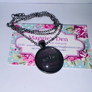 Peut inclure: Un collier pendentif noir avec un charme rond portant l'inscription "Oh you're good but I'm Crowley" en rose. Le collier repose sur une carte à motifs floraux avec le texte "Maggie's Den".