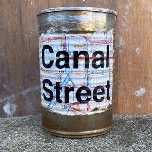May include: A rusty metal can with a map of the New York City subway system on it. The can has the words "Canal Street" printed on it in black.