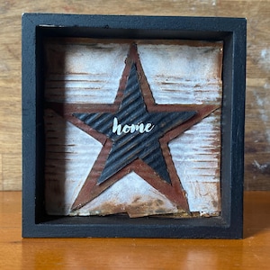 May include: A rustic, framed wall art piece featuring a star design. The star is made of corrugated metal and painted in black and red, with the word "home" in white script. The frame is black, and the background is white and brown.