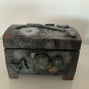 Könnte beinhalten: Eine kleine Holzkiste mit dunkelgrauer Oberfläche. Die Kiste ist mit Metallrädern, einem Schmetterling und einem strukturierten Design verziert. Die Kiste hat einen Klappdeckel und einen dekorativen Sockel.