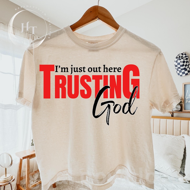 May include: A cream-colored t-shirt with the text "I'm just out here TRUSTING God." The word "TRUSTING" is in large red letters, while "God" is in a stylish black script. The shirt is hanging on a wooden hanger.