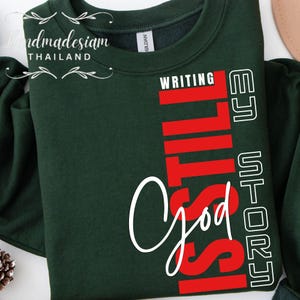 Może przedstawiać: Ciemnozielona bluza z napisem "WRITING IS STILL God's STORY" w kolorze białym i czerwonym. Bluza ma okrągły dekolt i długie rękawy. Marka "handmadesiam THAILAND" jest wydrukowana po lewej stronie u góry.